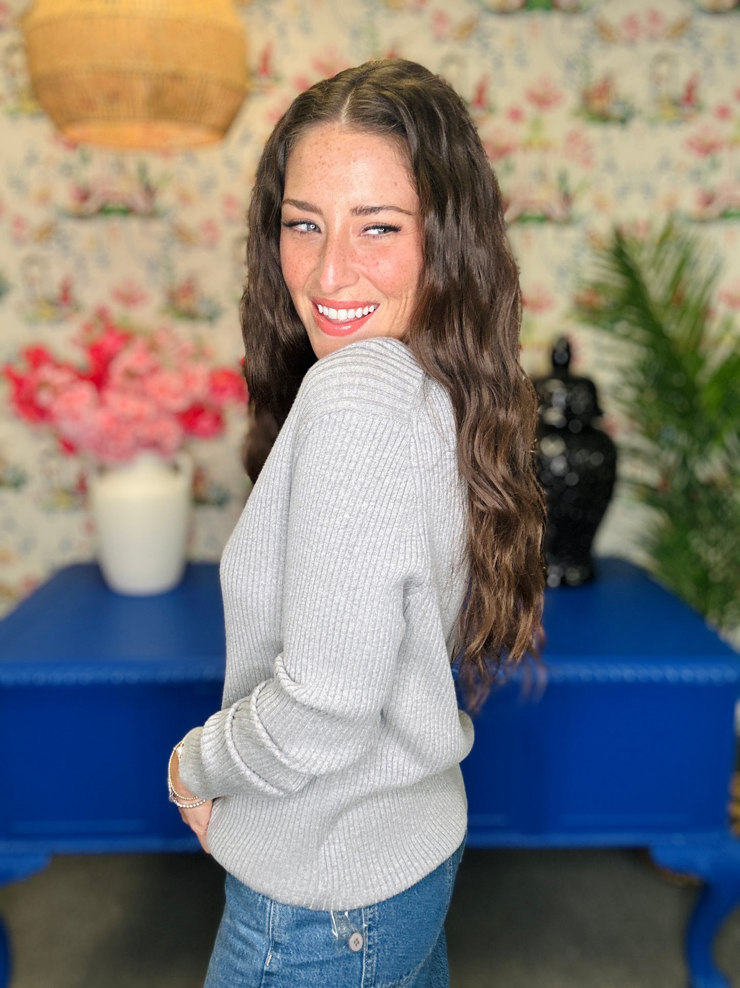 A woman with long wavy brown hair smiles while looking over her shoulder, wearing a light gray ribbed knit sweater and blue jeans in a decorated room with floral wallpaper and a blue table.