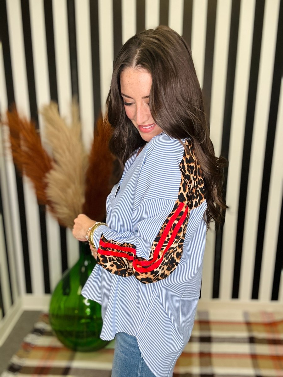 A woman wearing a light blue and white striped button-up shirt with leopard-print elbow patches and red piping details on the sleeves, paired with blue jeans, standing in front of a black and white striped wall.