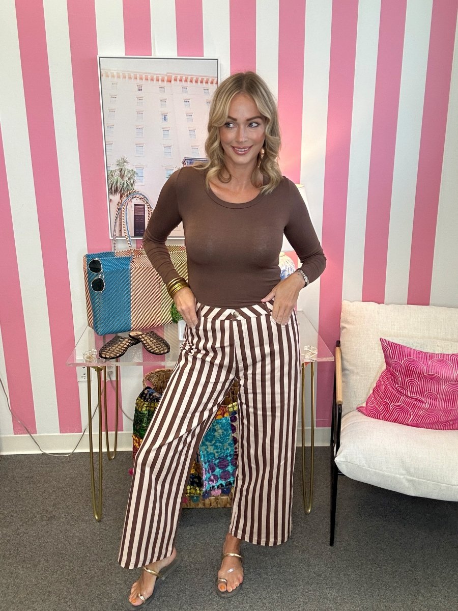 A woman stands in a room with pink and white striped wallpaper, wearing a brown long-sleeve top and brown-and-white vertically striped wide-leg pants. She is posing with her hands on her hips, standing next to a glass side table with a blue phone case and a white armchair with a pink pillow.