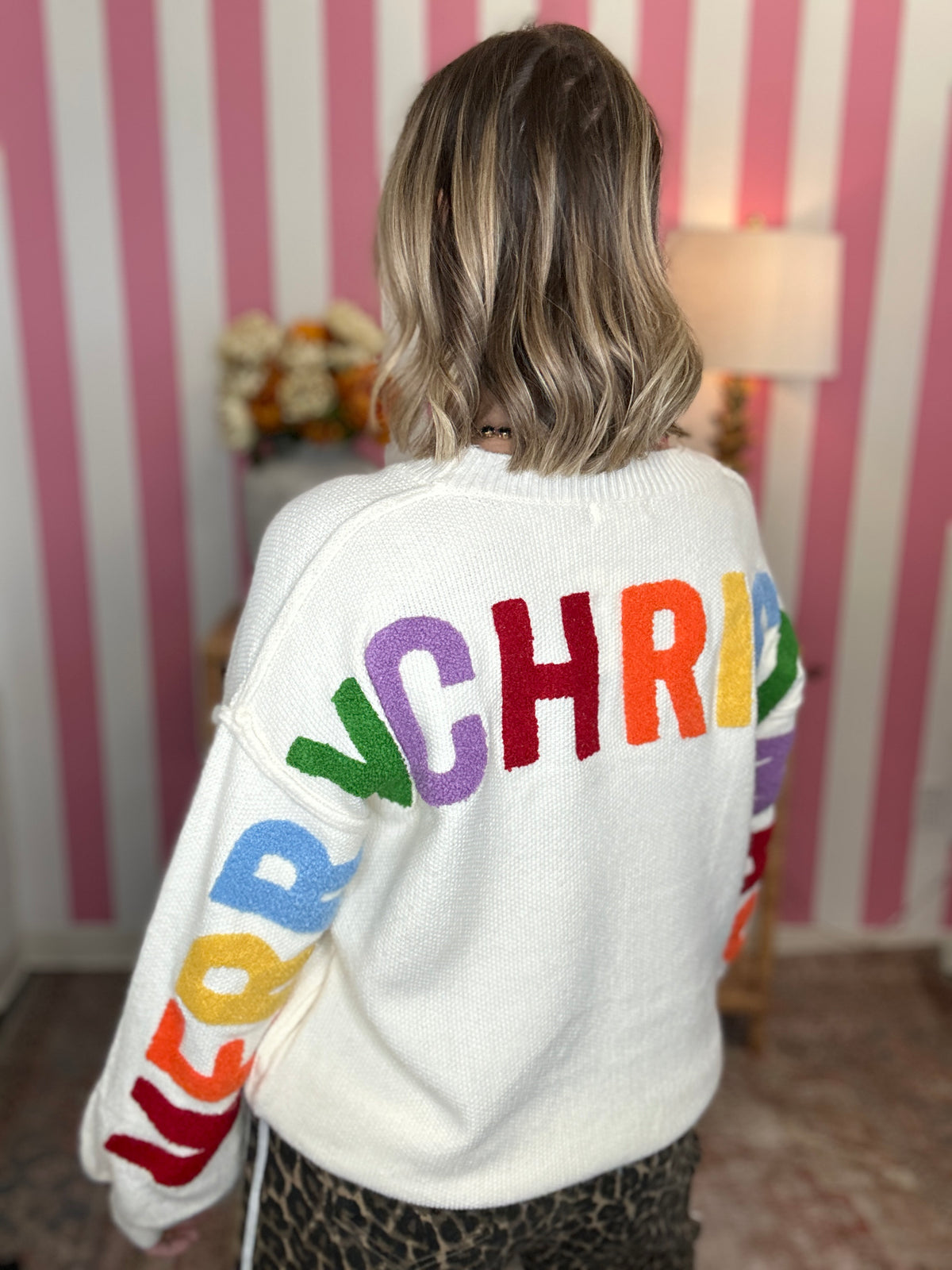 A woman viewed from behind wearing a white knit sweater with large multicolored letters spelling 'CHRIST' across the back and 'MARY' down one sleeve, set against a pink and white striped wall.