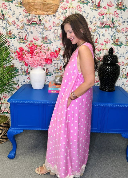 Woman in a pink polka dot dress standing in front of a blue table with decorative items and floral wallpaper.