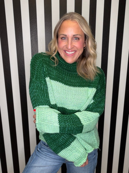 A smiling woman with blonde hair wearing a chunky knit sweater with green and mint green geometric blocks, paired with blue jeans, standing in front of a black and white striped wall.