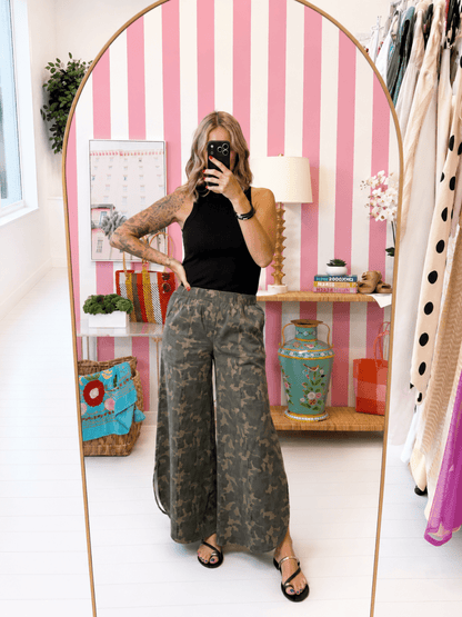Person taking a mirror selfie wearing a black top and camouflage pants in a room with pink striped wallpaper.