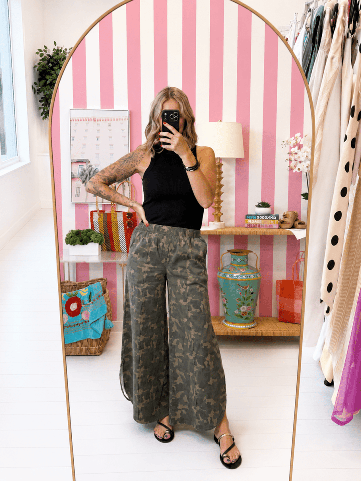 Person taking a mirror selfie wearing a black top and camouflage pants in a room with pink striped wallpaper.