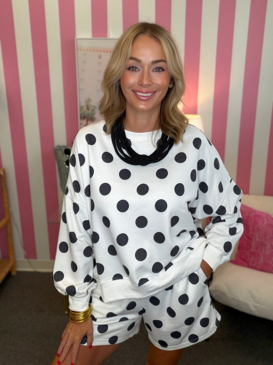 A woman wearing a white long-sleeve sweatshirt and matching shorts set with large black polka dots, standing in front of a pink and white striped wall.