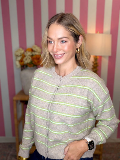 A woman wearing a light gray knit zip-up cardigan with thin lime green horizontal stripes, standing in front of a pink and white striped wall.