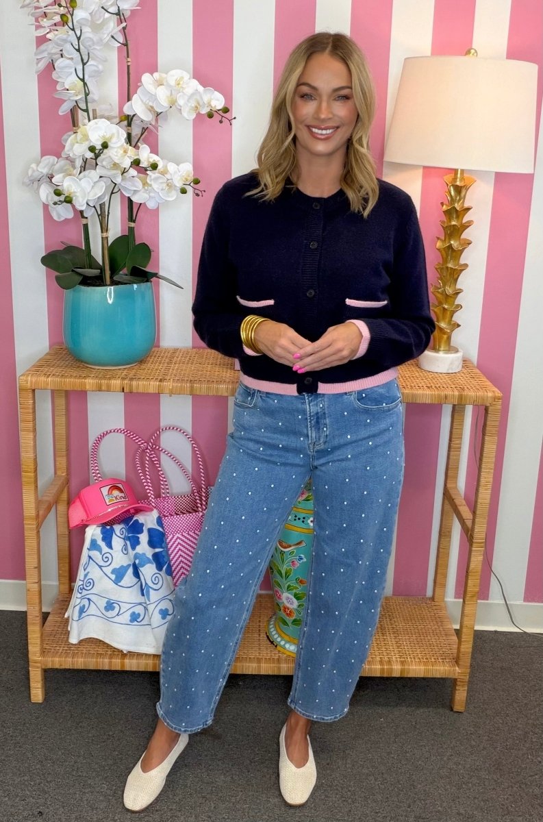 A woman stands in a styled room wearing a navy cardigan with pink trim and light blue jeans with white polka dots, posed in front of a pink and white striped wall with a wooden side table, lamp, and flowers.