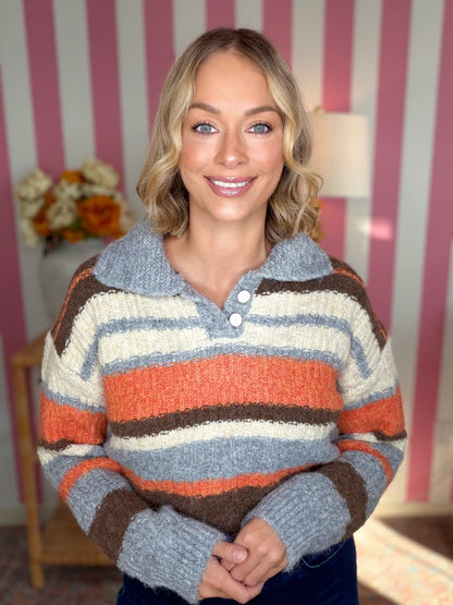 A smiling blonde woman wearing a fuzzy knit polo sweater with horizontal stripes in gray, orange, brown, and cream, posed in front of a pink and white striped wall.