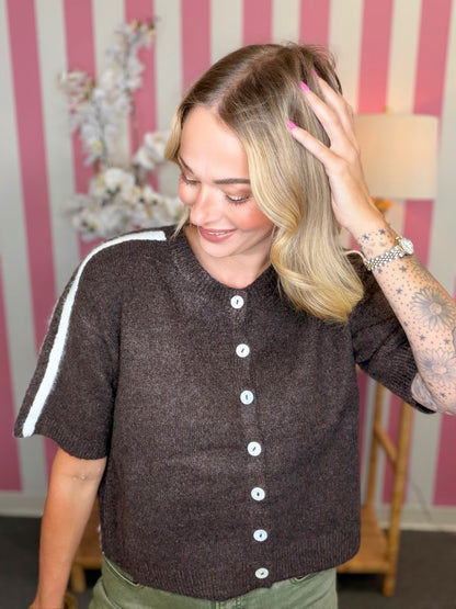 A woman wearing a dark brown short-sleeve knit cardigan with white buttons and a white stripe detail on the sleeve, standing in front of a pink and white striped wall.