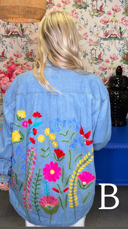 A woman viewed from behind wearing a light blue denim jacket with colorful embroidered wildflowers on the back, set against a patterned wallpaper background.