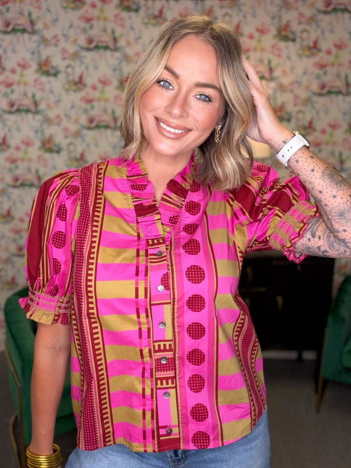 A woman wearing a vibrant pink and gold striped blouse with bohemian-style patterns, posing in front of a floral wallpaper background.