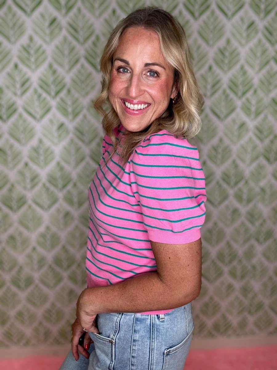 A smiling blonde woman wearing a pink short-sleeve polo shirt with thin blue horizontal stripes and light-wash blue jeans, standing in front of a green and white patterned wall.