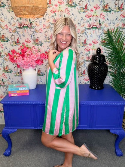 A woman posing in a vibrant green and pink vertically striped tunic dress, standing in front of a blue side table decorated with a vase of pink flowers and a black ceramic jar against a patterned wallpaper background.