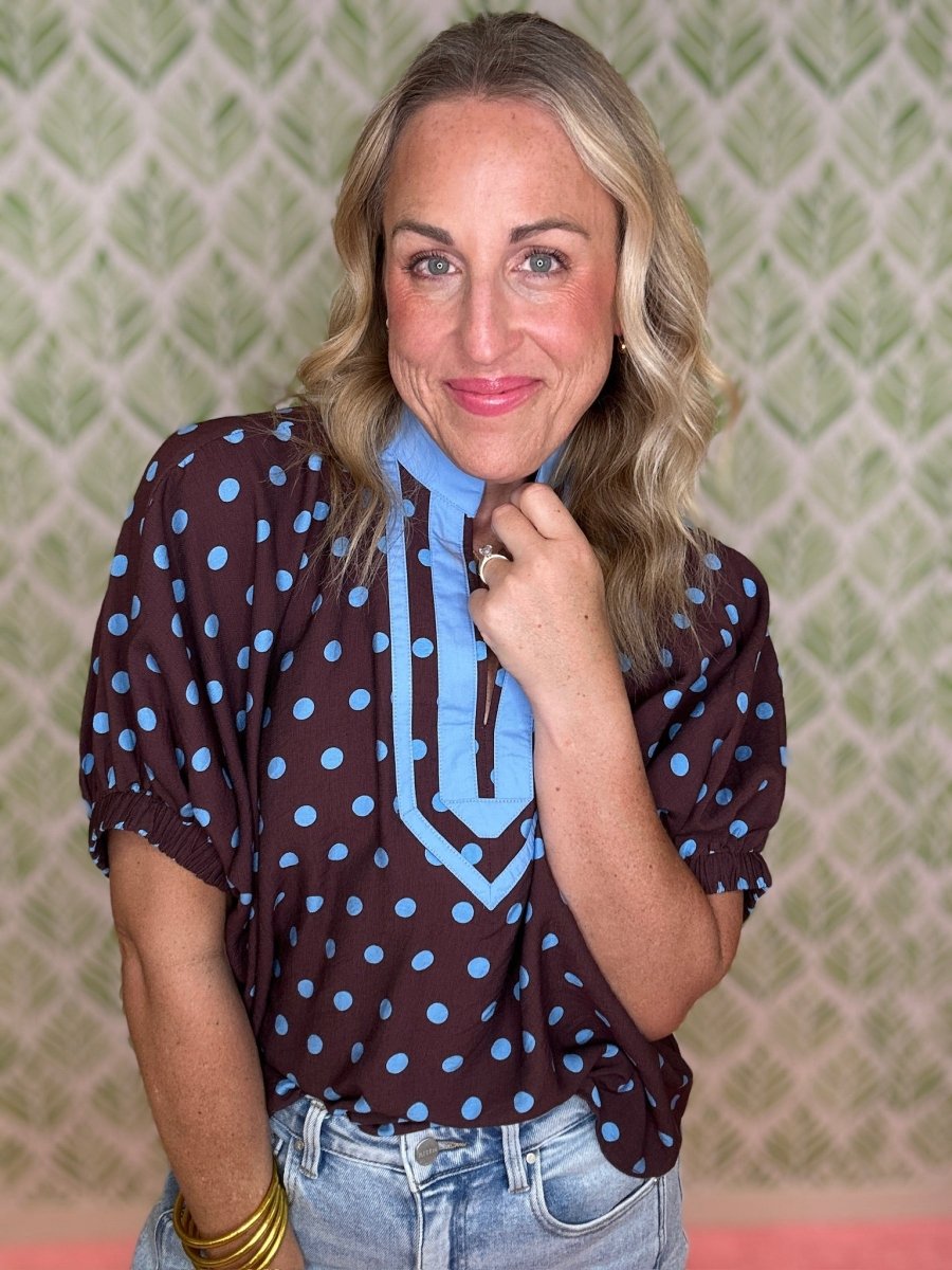 A woman wearing a brown short-sleeve blouse with light blue polka dots and a light blue tie-neck detail, paired with light-wash blue jeans, posing in front of a green patterned wallpaper background.