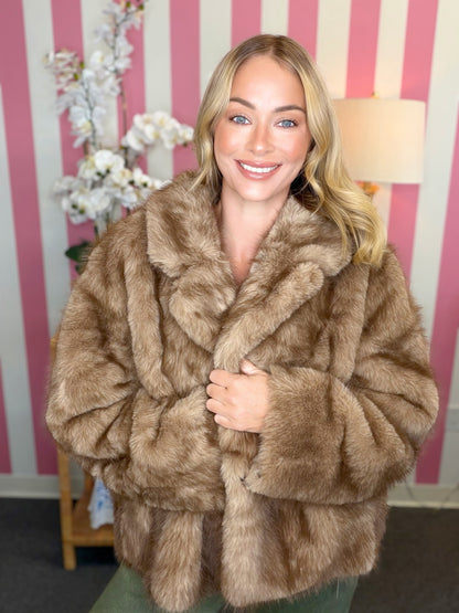 A smiling blonde woman wearing a voluminous brown faux fur coat, standing in front of a pink and white striped wall.
