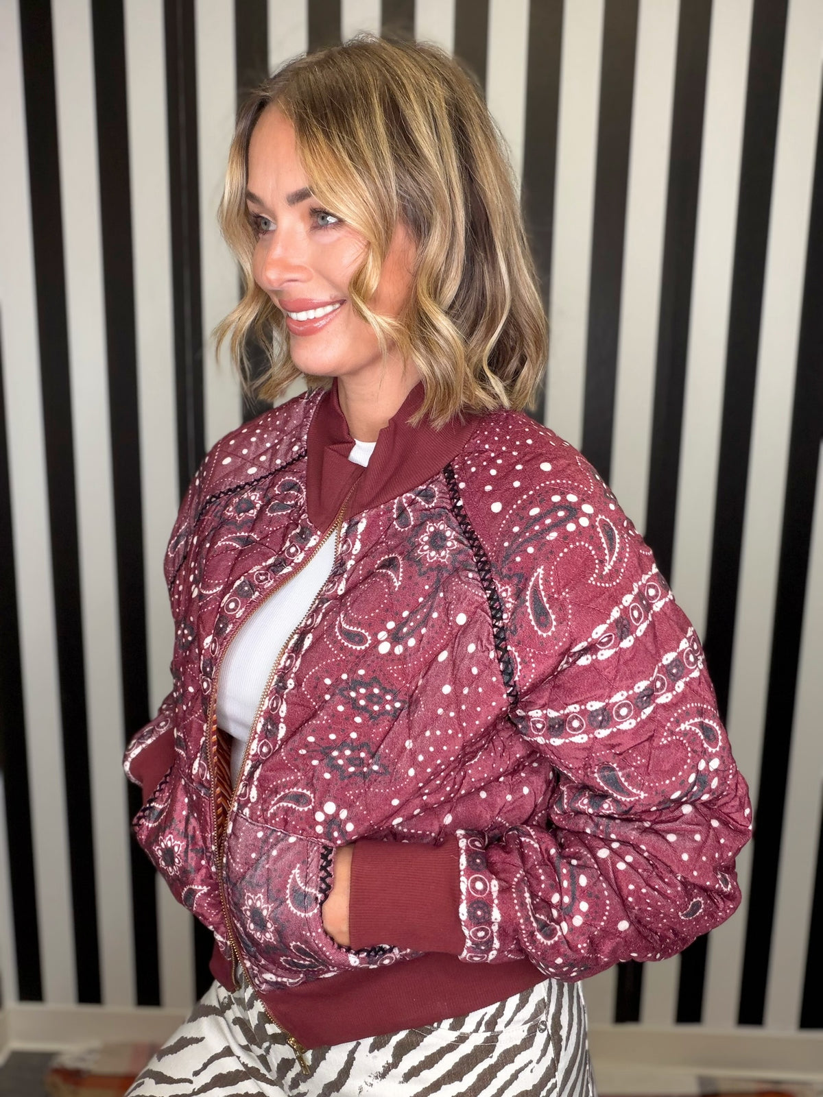 A woman wearing a burgundy and black paisley bandana bomber jacket over a white top, standing in front of a black and white striped wall.