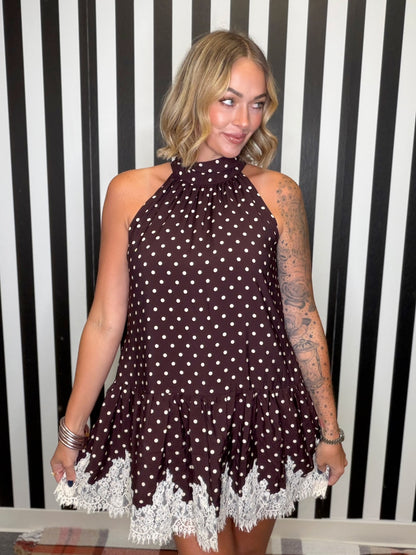 A woman wearing a sleeveless halter-neck mini dress with a brown and white polka dot pattern and a white lace hem, standing in front of a black and white striped wall.