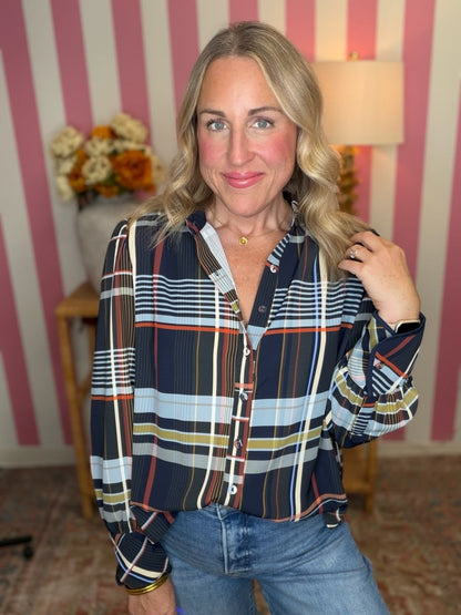 A woman wearing a long-sleeve button-up plaid shirt with navy, blue, white, yellow, and red stripes, paired with blue jeans, standing in front of pink and white striped wallpaper.