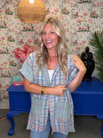 A woman with blonde hair stands indoors wearing a light blue and white plaid short-sleeve button-front vest over a white top and jeans. The background features floral wallpaper, a blue table, and a woven pendant light.