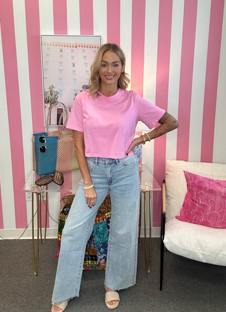 A woman posing in a pink short-sleeve crop top and light-wash wide-leg jeans, standing in front of a pink and white striped wall with handbags and a chair.