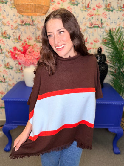 A woman wearing a brown knit poncho sweater with a wide light blue stripe and red borders, standing in front of a floral wallpaper background with a blue side table.