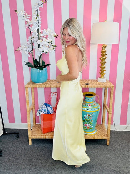 Woman in a yellow dress standing next to a decorative table with vases and a lamp against a pink and white striped wall.