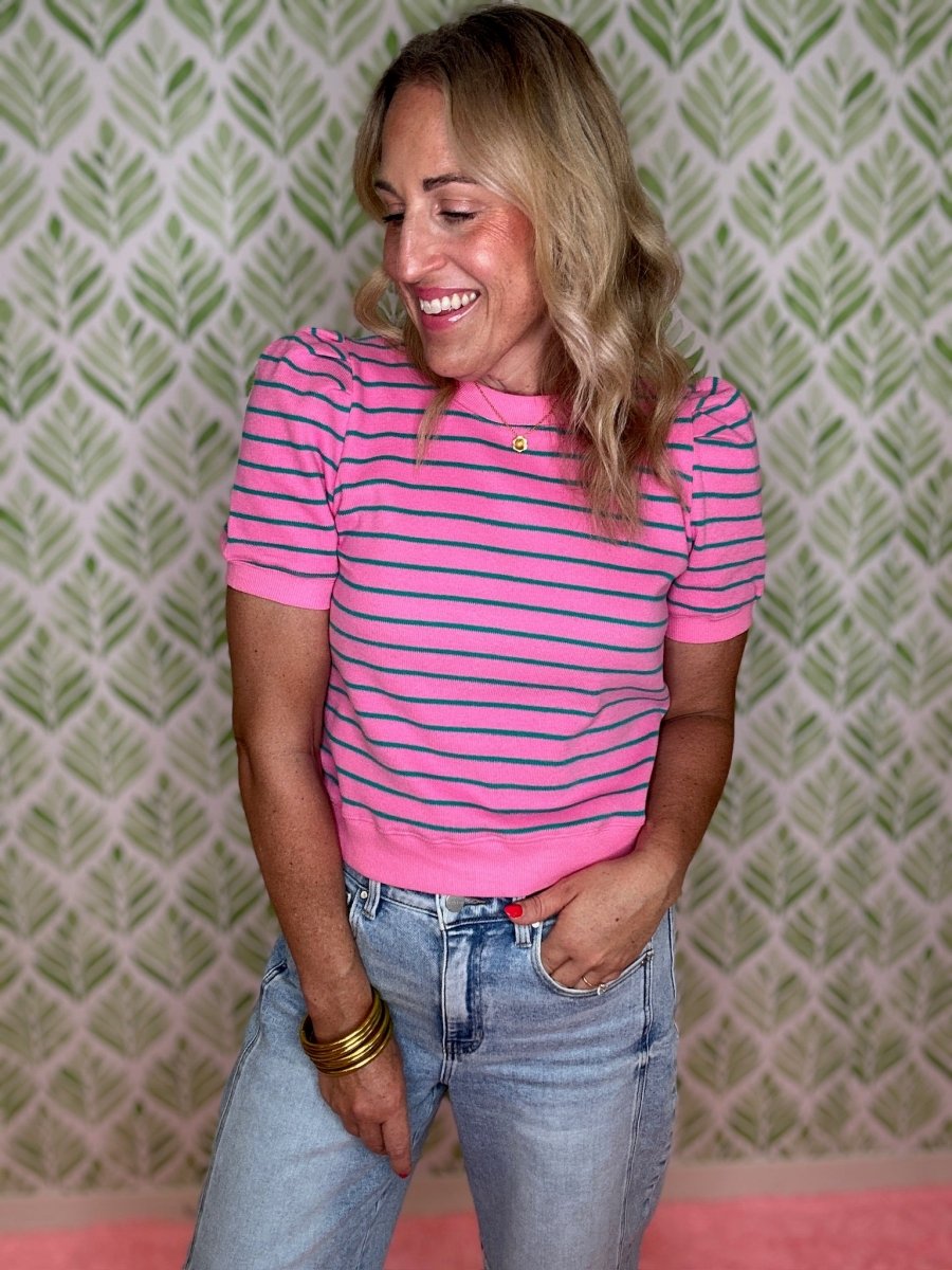 A woman wearing a pink short-sleeve striped knit top with blue jeans, standing in front of a green and white patterned wallpaper background.
