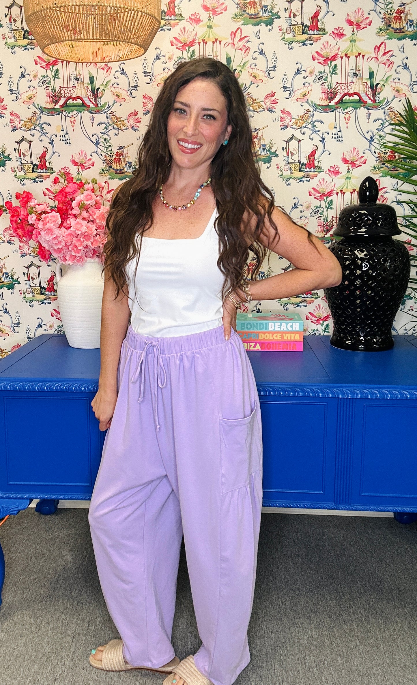 A woman stands indoors wearing a white sleeveless top and loose lavender drawstring pants, posed in front of a floral wallpaper wall and a blue cabinet.