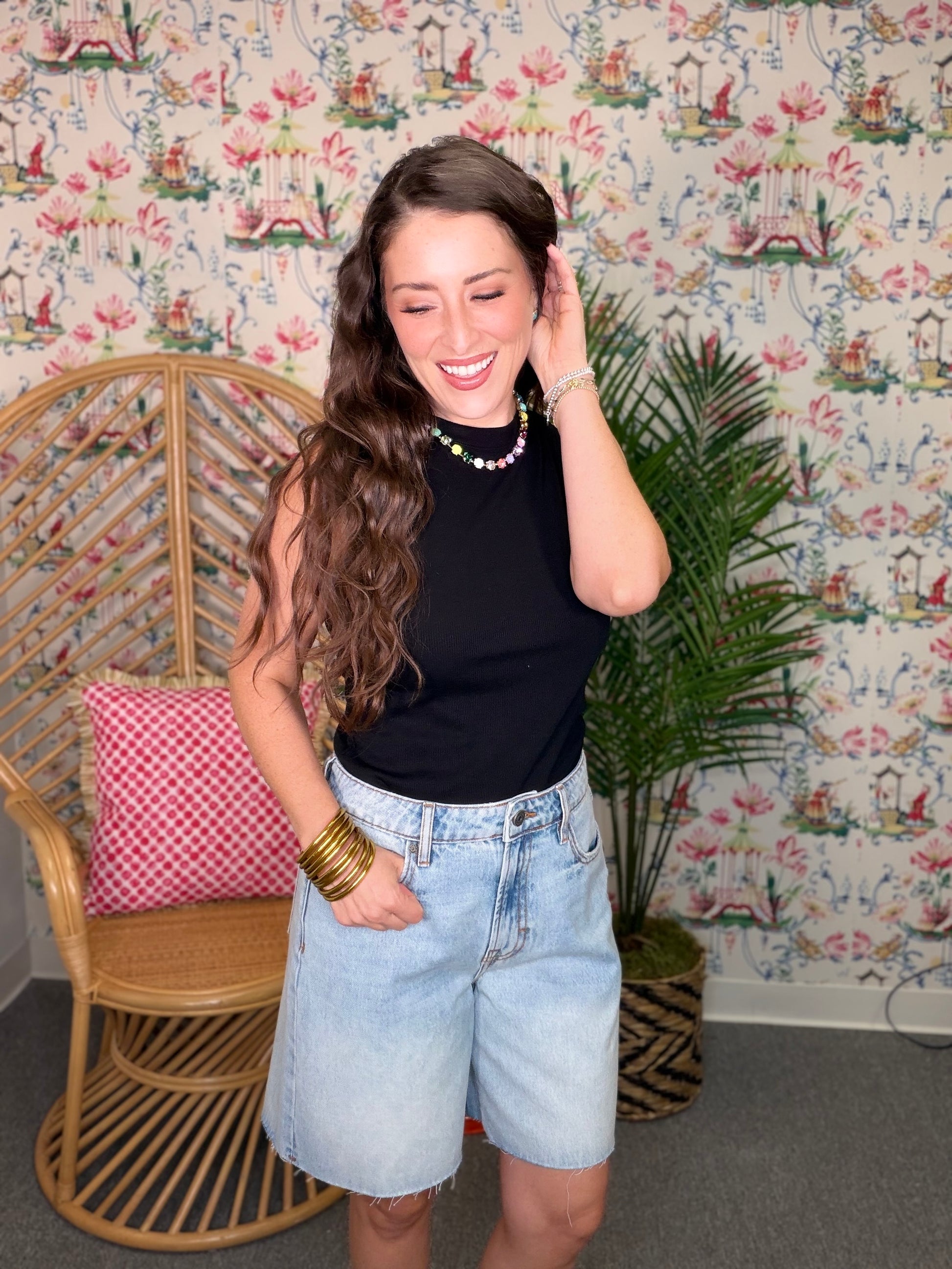 Woman in black sleeveless top and denim shorts standing in a room with floral wallpaper.