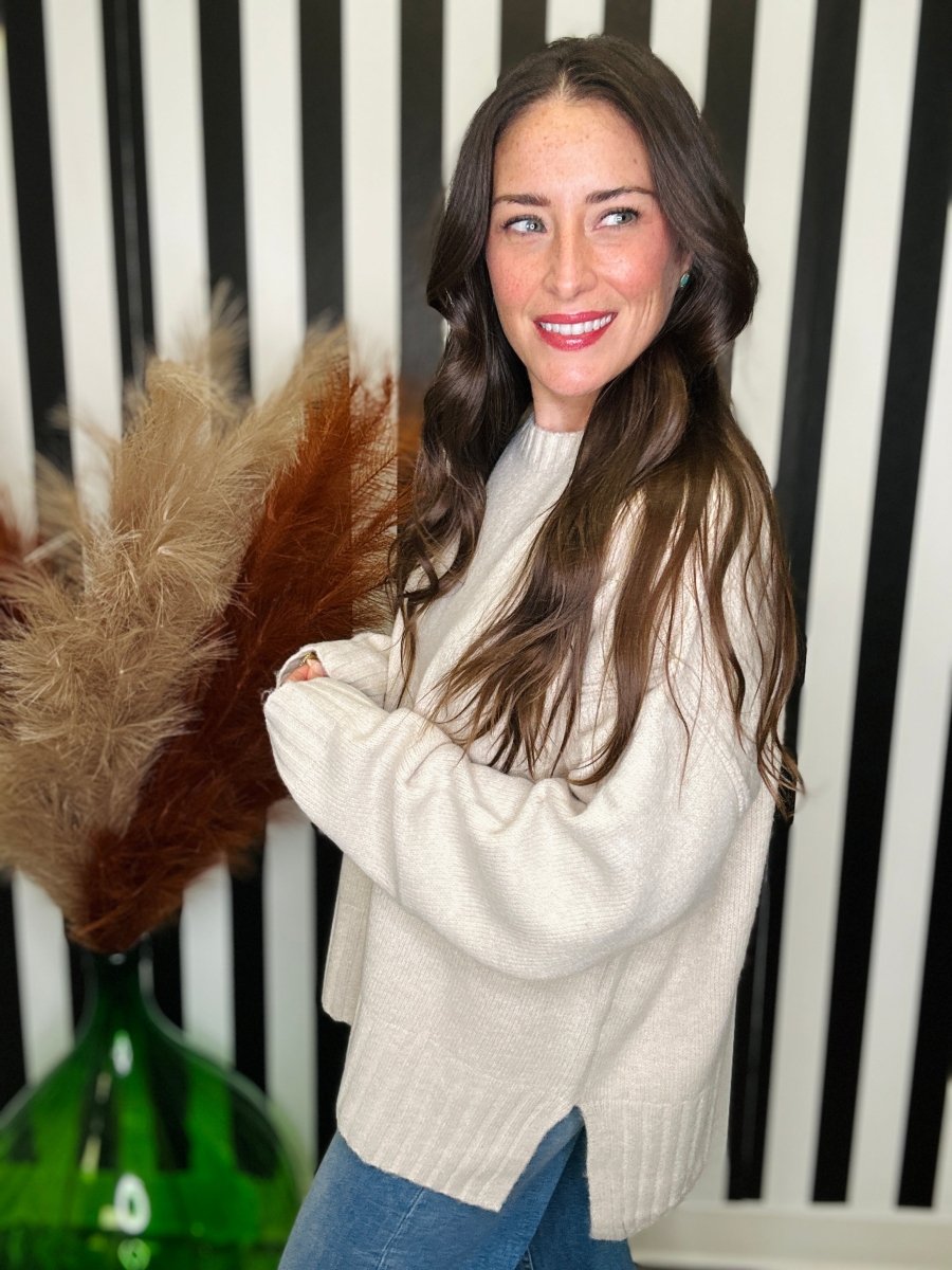 A woman with long brown hair wearing a cream-colored knit sweater and blue jeans, standing in front of a black and white striped wall with dried pampas grass in a green vase.