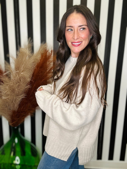 A woman with long brown hair wearing a cream-colored knit sweater and blue jeans, standing in front of a black and white striped wall with dried pampas grass in a green vase.