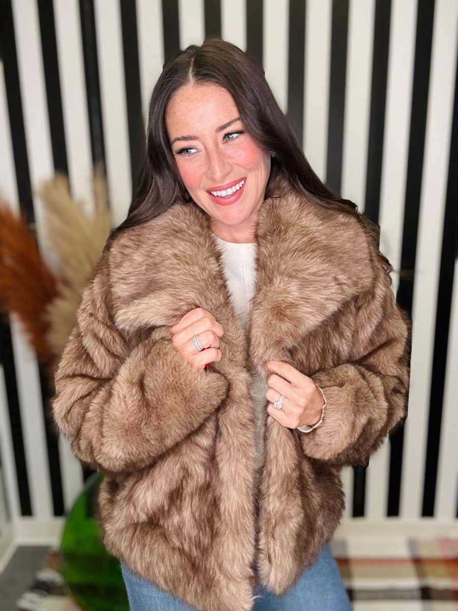 A woman wearing a plush, light brown faux fur jacket over a white top and blue jeans, standing in front of a black and white striped wall.