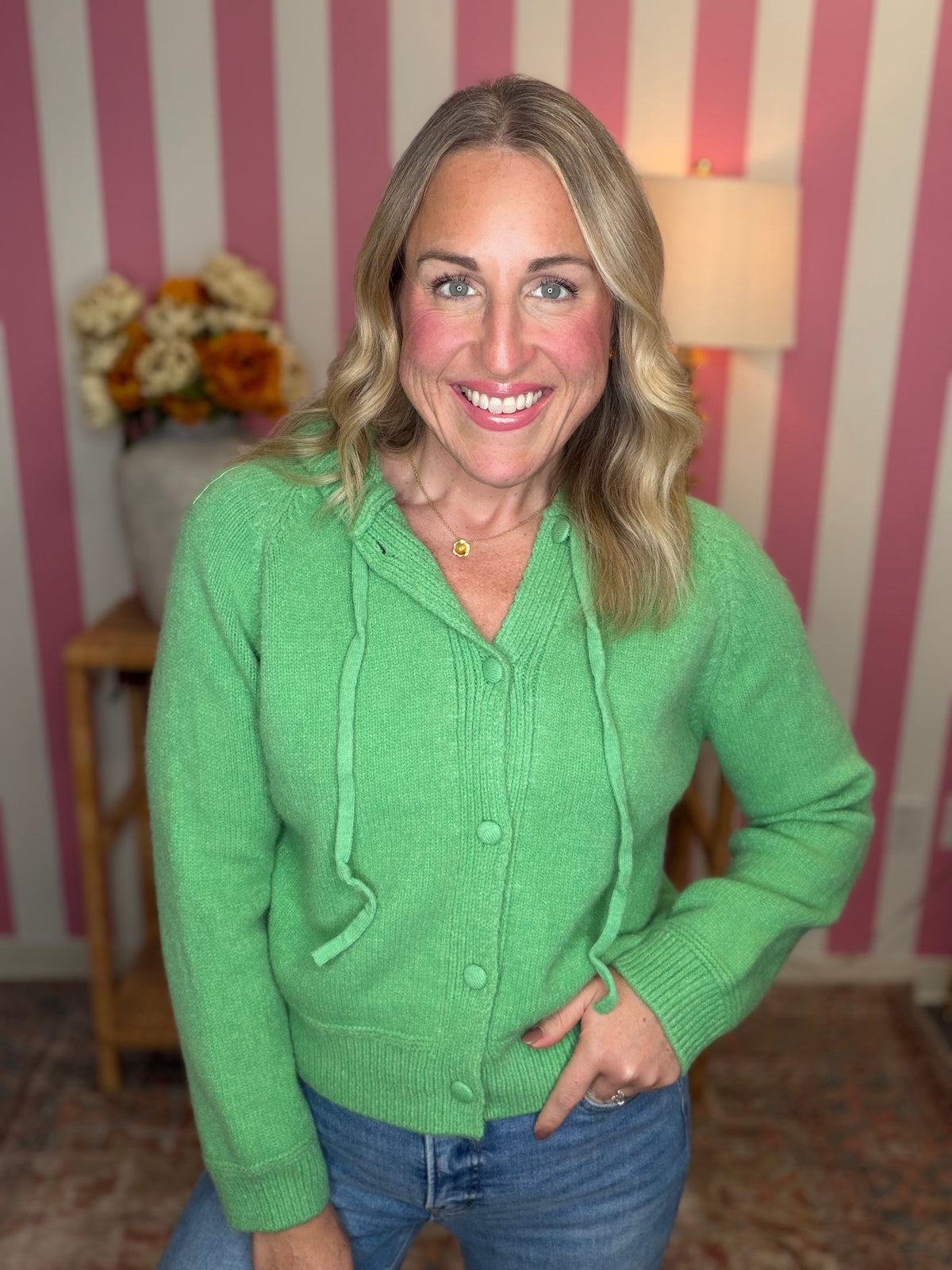 A smiling blonde woman wearing a bright green knit cardigan with drawstrings and buttons, paired with blue jeans, standing in front of a pink and white striped wall.