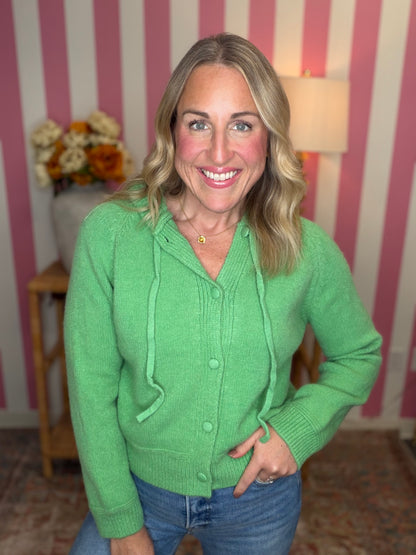 A smiling blonde woman wearing a bright green knit cardigan with drawstrings and buttons, paired with blue jeans, standing in front of a pink and white striped wall.