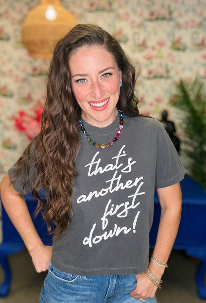 A smiling woman with long wavy brown hair wearing a dark gray graphic t-shirt with the white text 'that's another first down!' and blue jeans, standing in a room with a floral wallpaper background.