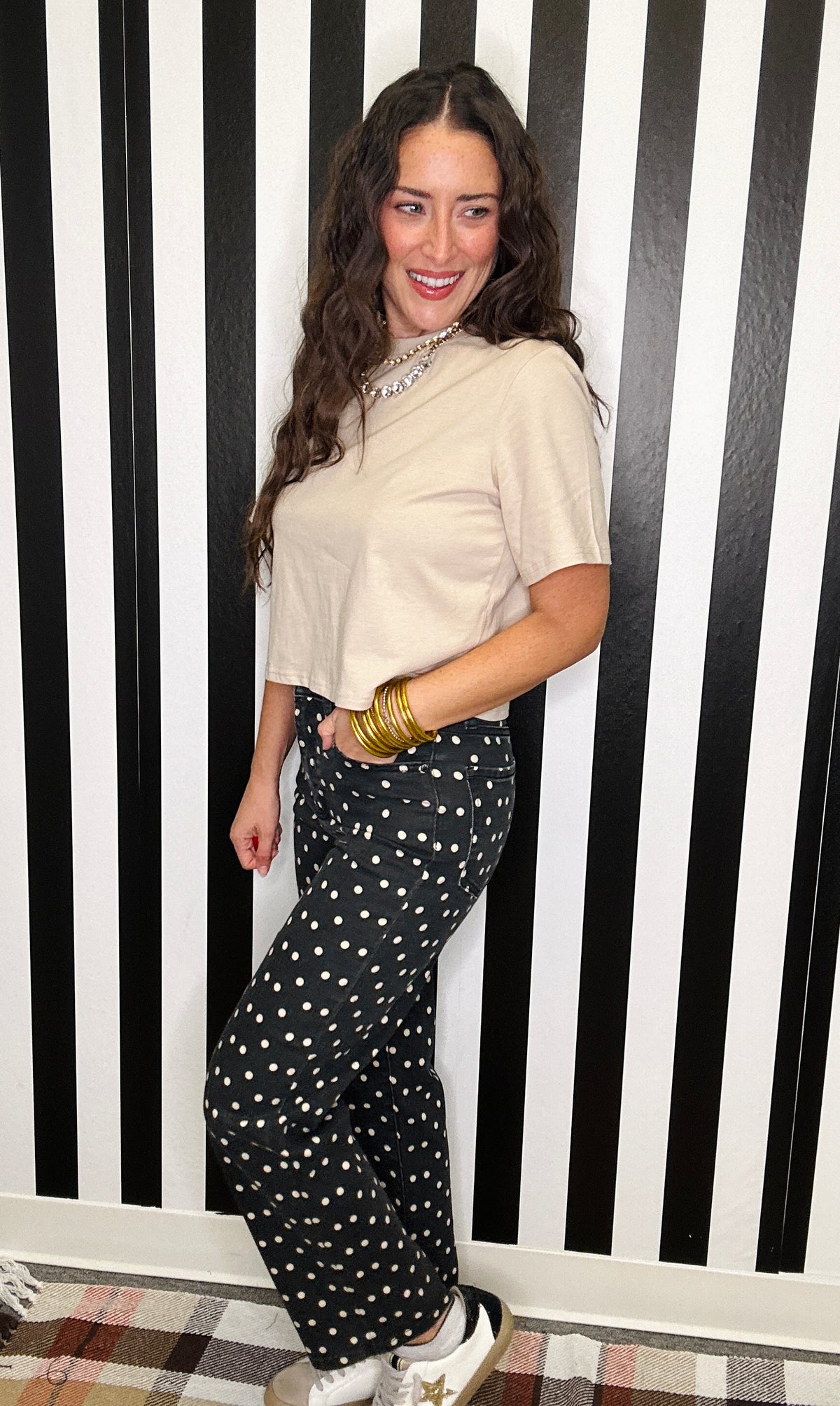 A woman posing in front of a black and white striped wall, wearing a beige short-sleeve top, black wide-leg pants with white polka dots, and white sneakers.