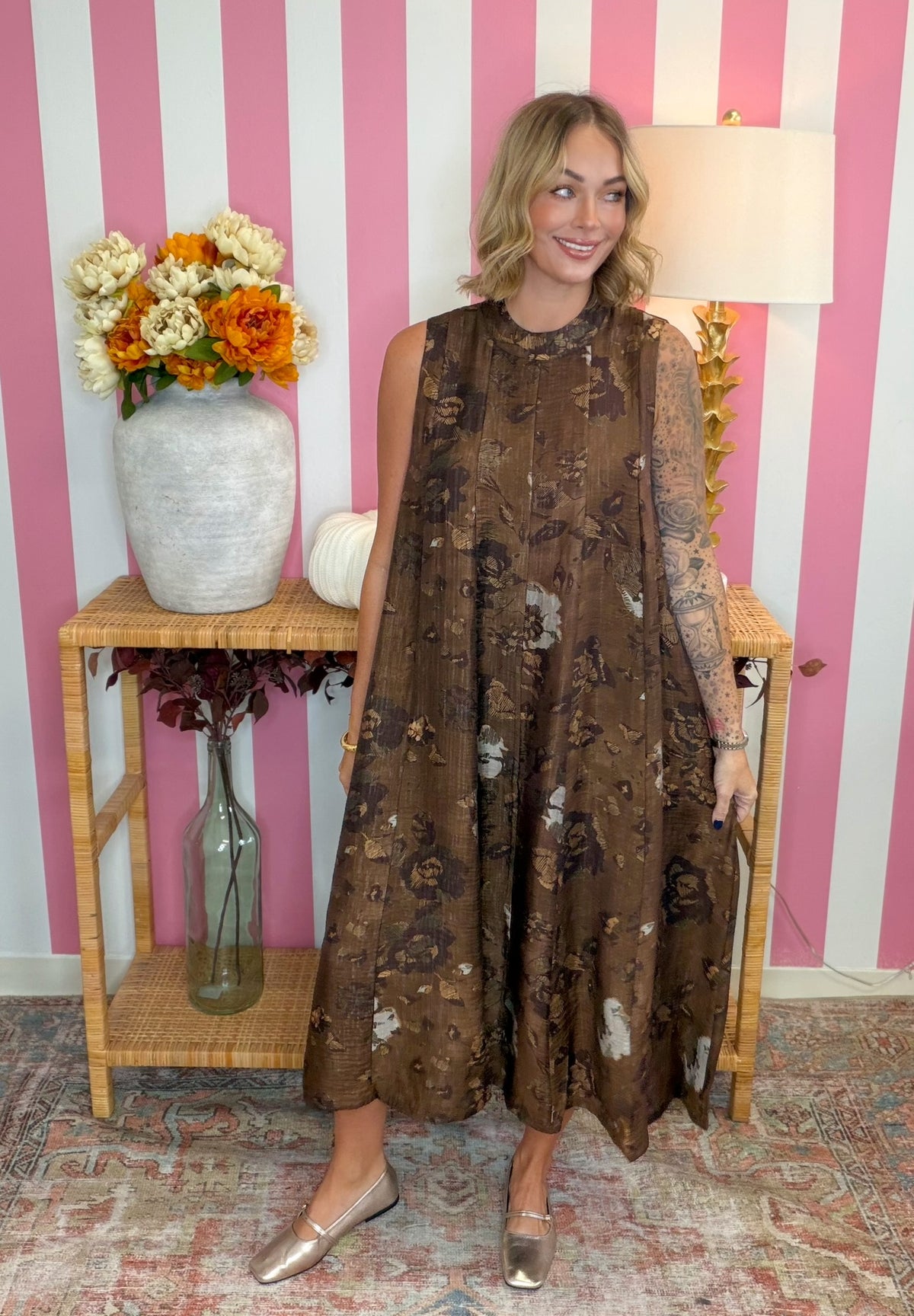 A woman modeling a brown sleeveless midi dress with a floral and abstract print, standing in front of a pink and white striped wall with a wooden side table and flowers.