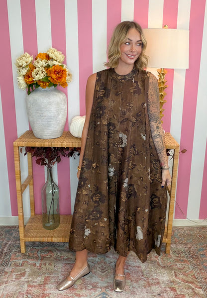 A woman modeling a brown sleeveless midi dress with a floral and abstract print, standing in front of a pink and white striped wall with a wooden side table and flowers.