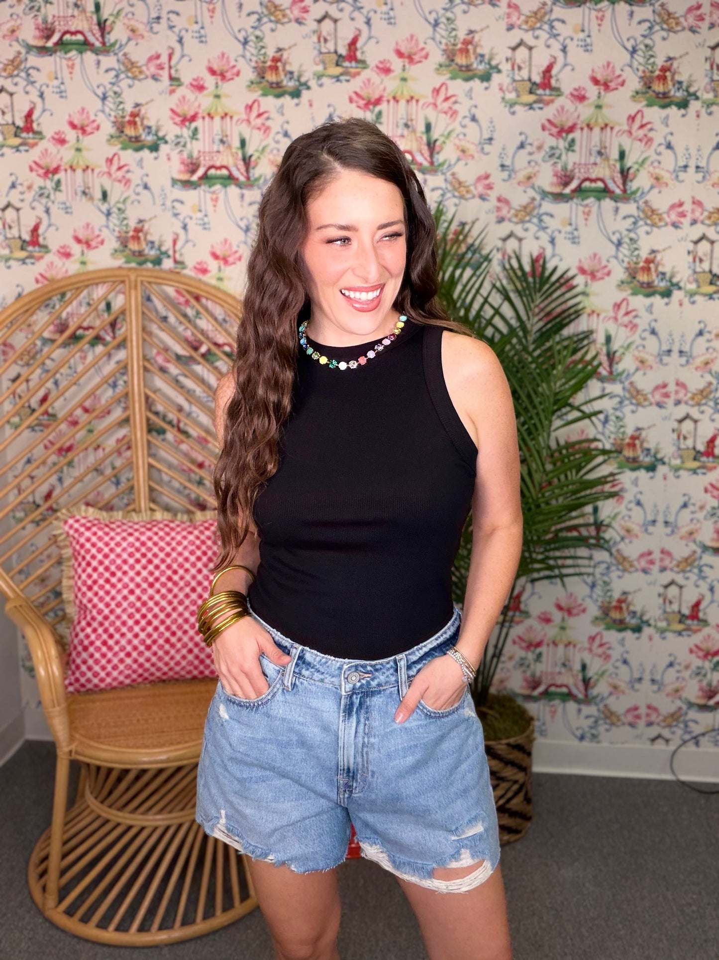 Woman wearing a black sleeveless top and denim shorts standing in a room with floral wallpaper.