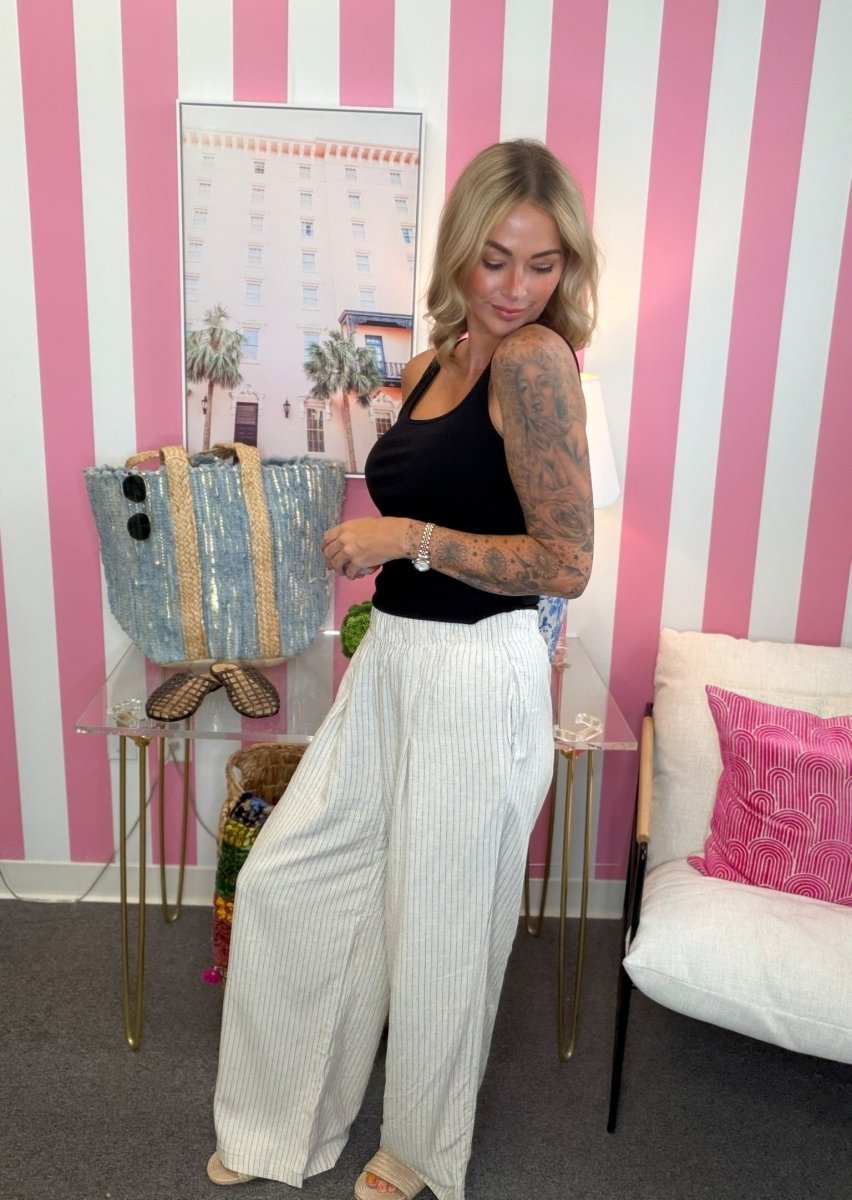 A woman stands in a room with pink and white striped wallpaper, wearing a black tank top and white pinstriped wide-leg trousers. She is holding a light blue sequined tote bag on a glass table next to a white sofa with a pink pillow.