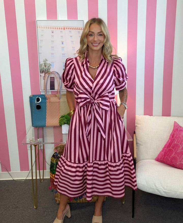 A woman wearing a pink and white vertically striped midi dress with puff sleeves and a self-tie waist, standing in front of a pink and white striped wall.