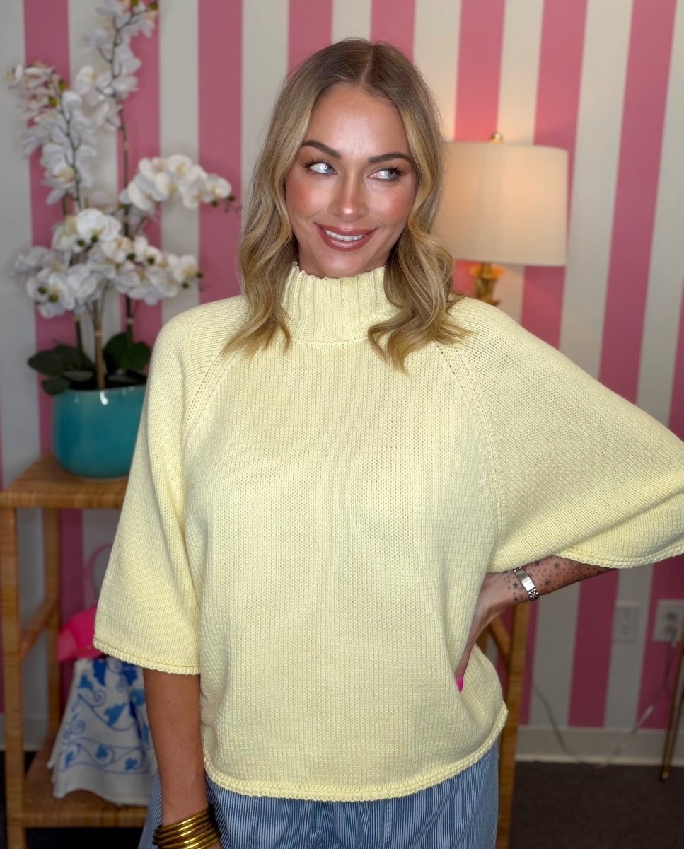 A woman wearing a pale yellow knit mock-neck sweater with wide three-quarter sleeves, standing in front of a pink and white striped wall.
