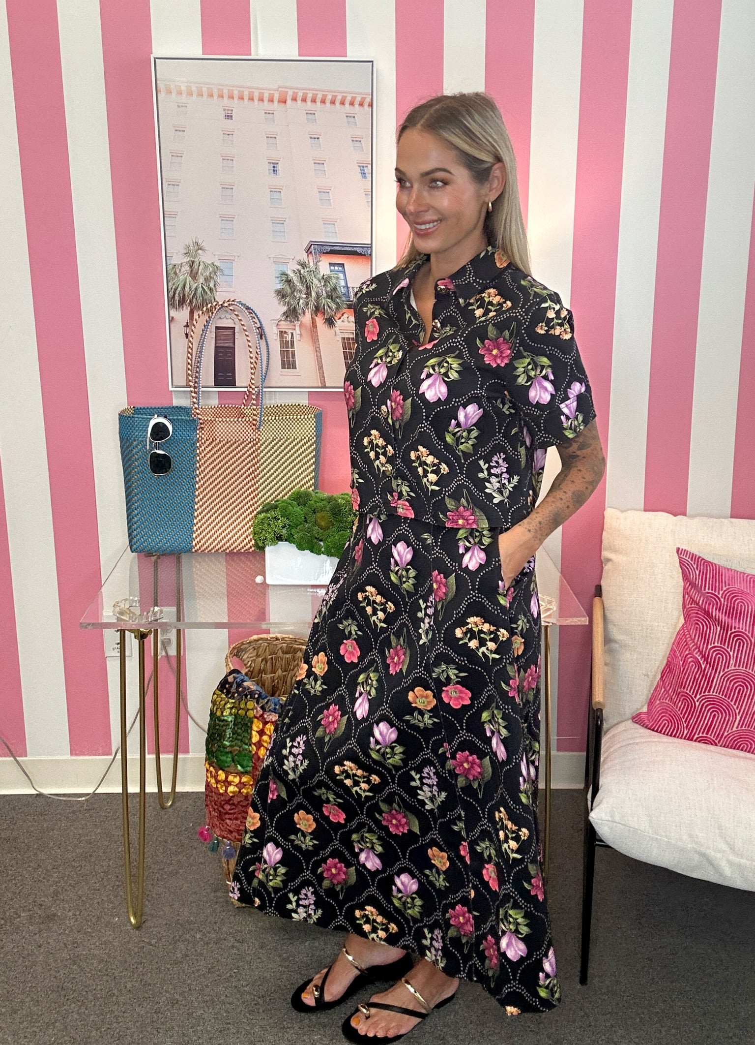 A woman wearing a black short-sleeve maxi dress with a colorful floral print stands in a room with pink and white striped walls. She is wearing black sandals and has her hand in her pocket.