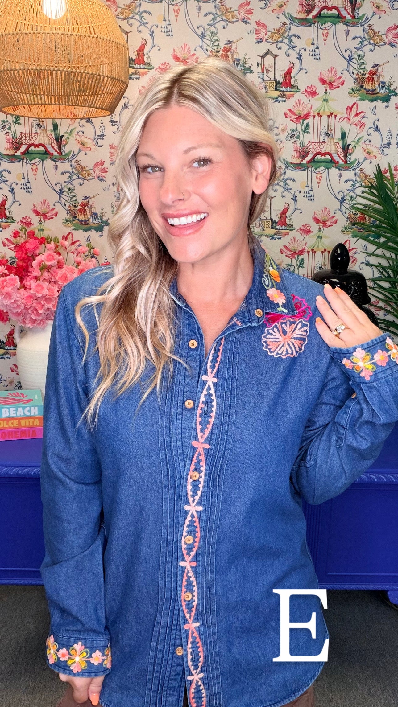 A smiling blonde woman wearing a blue denim button-up shirt with colorful embroidered details on the front placket, cuffs, and chest, posed in front of a floral wallpaper background.