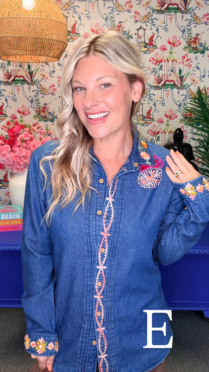 A smiling blonde woman wearing a blue denim button-up shirt with colorful embroidered details on the front placket, cuffs, and chest, posed in front of a floral wallpaper background.