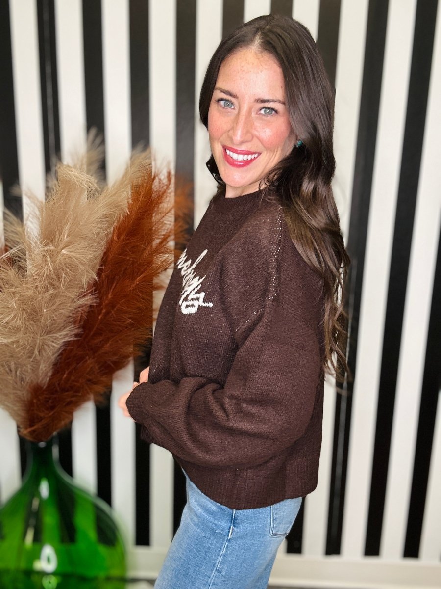 A woman wearing a brown knit sweater with white script embroidery on the chest, paired with light blue jeans, standing in front of a black and white striped background.