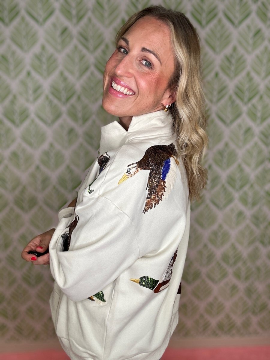 A smiling blonde woman wearing a white high-neck pullover sweatshirt with detailed duck hunting graphics, including a large duck wing on the shoulder and a duck on the pocket, posed against a green patterned wallpaper background.