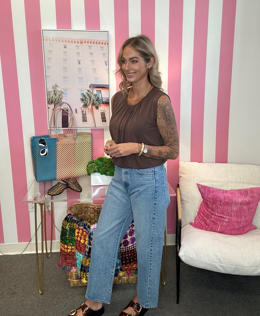 A woman stands in a room with pink and white striped walls, wearing a brown sleeveless top, light blue wide-leg jeans, and black sandals. She is posing next to a glass table displaying colorful handbags and a white armchair with a pink pillow.