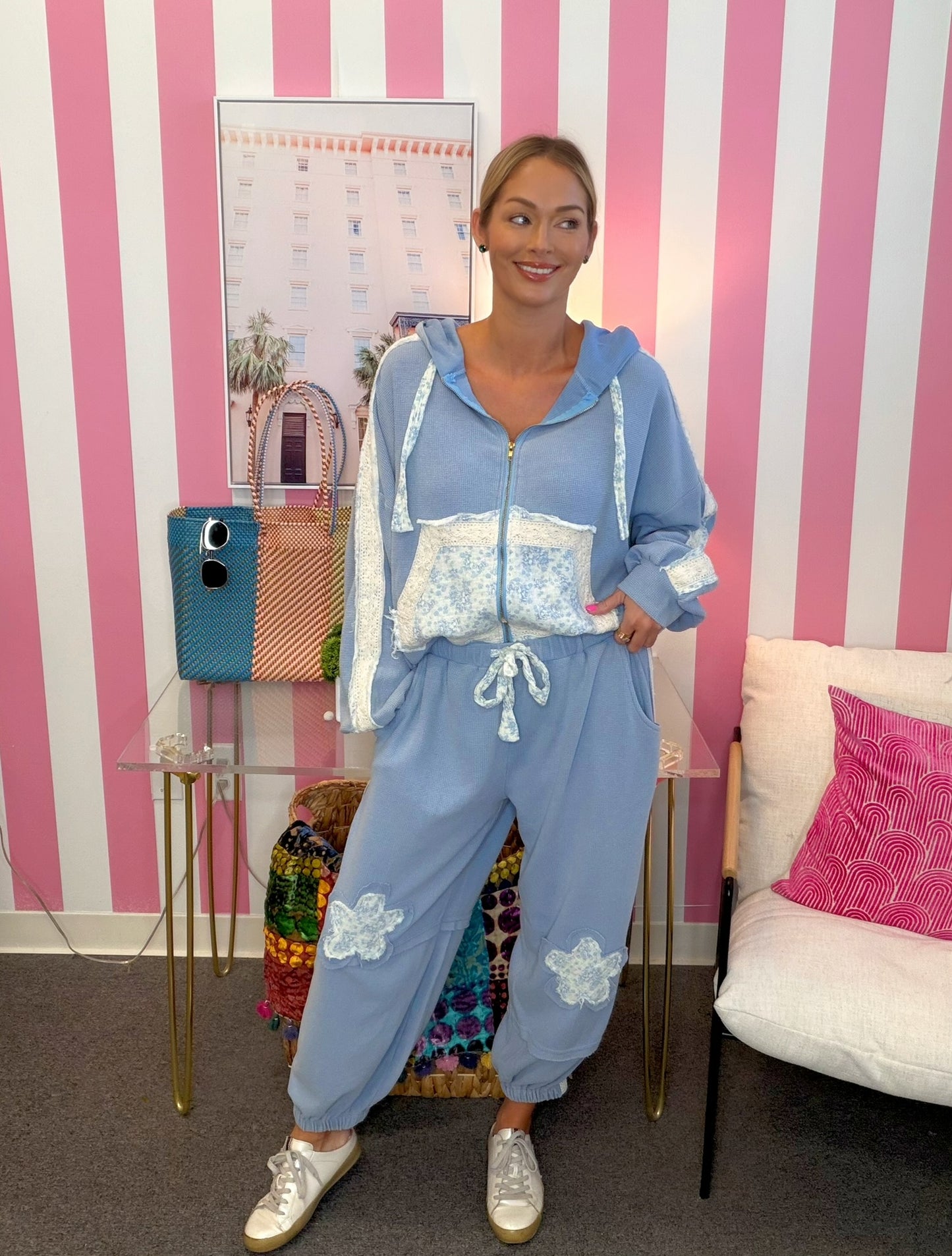 A woman modeling a light blue zip-up hoodie and matching jogger pants set with white lace details, standing in front of a pink and white striped wall.