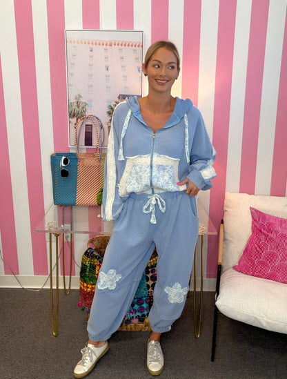 A woman modeling a light blue zip-up hoodie and matching jogger pants set with white lace details, standing in front of a pink and white striped wall.